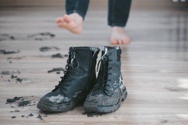 Mud black boots next to muddy footprints and major mud cakes on a wooden floor in living room. Bare feet of a person on the background. Concept of dirty stains and house cleaning. . High quality photo clipart