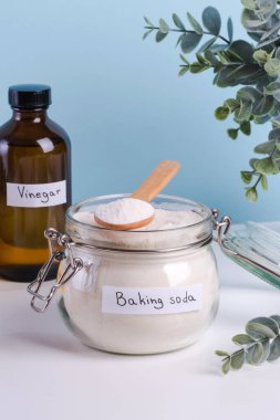 Kabartma tozu ve ahşap kaşıkla dolu bir kavanoz mavi arka planda bir şişe sirkeyle birlikte tezgahın üzerinde duruyor. Lekeleri çıkarmak için doğal ürünleri kullanarak DIY temizlik yöntemi.
