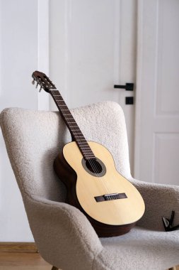İyi işlenmiş bir akustik gitar parlak, davetkar bir oturma odasında yumuşak, krem renkli bir sandalyeye yaslanır. Minimalist dekor sakin bir atmosfer yaratıyor. Yüksek kalite fotoğraf