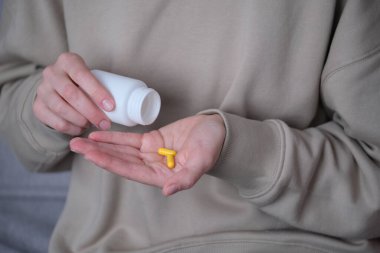 Bir insan evde rahat bir şekilde otururken beyaz bir şişeden sarı kapsülleri dikkatlice bir eline döker. Yumuşak ışıklandırma anın detaylarını vurguluyor. Yüksek kalite fotoğraf