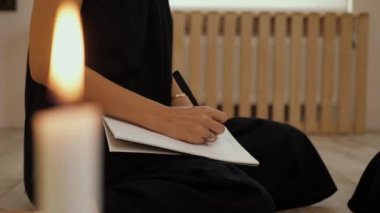 Siyahlı genç bir kadının eli deftere yazıyor, ödevlerini yapıyor, ön planda mum var. Yaşam tarzı ve evde çalışma kavramı. Kadın yazar eli, yakın plan.