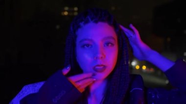 Neon moda Asyalı kadın gece caddesinde yüzüne yavaşça dokunuyor, yüzeysel odaklanma. Hippi eğilimi Vaporwave. Çok renkli kırmızı ve mavi renkli genç Japon kadın kameraya bakıyor.