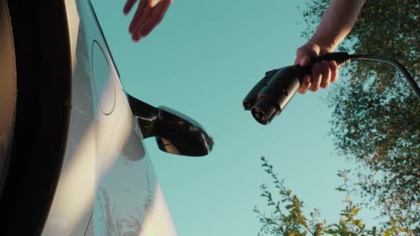 男性の手はEv車 ローアングル クローズアップに電源コネクタを挿入します 認識できない男は電気自動車に充電ケーブルを接続し 青空と緑の木の背景にバッテリーを充電します — ストック動画