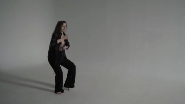 Siyah elbiseli güzel kıvırcık kadın, beyaz bir fotoğraf stüdyosunda, bir siklonamada yavaşça yalınayak dans ediyor.