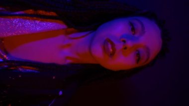 Çok renkli, mavi ışık saçan, kameraya bakan, sığ odaklı havalı genç Asyalı kadının dikey videosu. Şehir tarzı. Neon Moda Hipster eğilimi