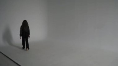 Siyah elbiseli güzel kıvırcık kadın beyaz bir fotoğraf stüdyosunun köşesinde çıplak ayakla dans ediyor.