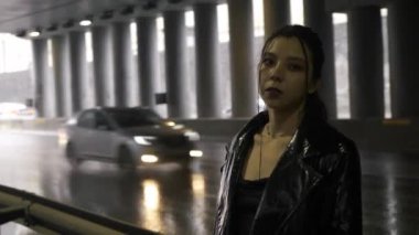 Köprünün altında siyah ceketli bir kadın kameraya bakıyor. Asyalı kadın arabalı bir yolun arka planında duruyor.