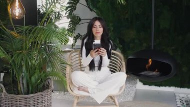 Beyaz elbiseli, elinde telefon olan güzel bir kadın, yeşil ve şömineli bir sandalyede oturuyor, evde dinleniyor, akıllı telefondan arkadaşlarıyla sohbet ediyor.