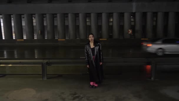 在桥下穿着黑色浅色外套的亚洲女歌手 年轻女子唱着歌 站在一条有车的大路的后面 看着摄像机 — 图库视频影像