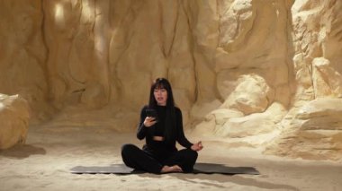 Siyah elbiseli güzel bir kadın kumda, çöl kumunda bacak bacak üstüne atarak oturur. Telefonla meditasyon yapar. İç uyumu hisseder.