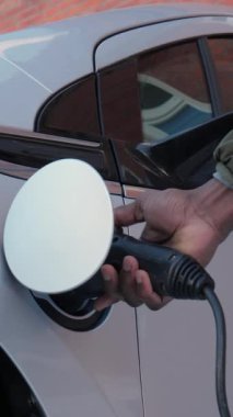 Tanımlanamayan Afrikalı adamın dikey videosu elektrik kablosunu elektrikli araçtan ayırdı. Afrikalı erkek el, EV 'den eski bir tuğla duvarın yakınındaki güç bağlantısını kesiyor. Şarj pillerini bitir