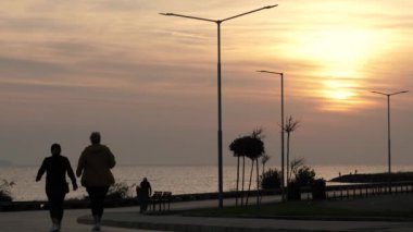 Şafak vakti deniz kenarındaki toprak yol. Tanınmayan insanlar deniz kenarında yürür.