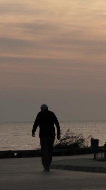 Denizin kenarında yürüyen, uzayı kopyalayan, tanınamayan yaşlı bir insanın dikey videosu. Şafak vakti deniz kenarındaki toprak yol.