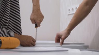 Modern bir evde, tanınmayan iki birey mobilyaları bir araya getirmek, takım çalışmasını ve becerilerini ön plana çıkarmak için birlikte çalışırlar. Tornavida, onarımın verimliliğini vurgulayarak DIY yaklaşımını yansıtır.