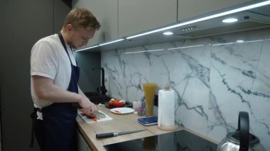 Parlak ışıklandırması olan modern bir mutfakta, önlüklü bir adam taze sebzeleri doğruyor. Şık mermer kaplama, sağlıklı bir yemek hazırlarken sofistike bir zemin hazırlar ve yemek pişirme tutkusunu gösterir.