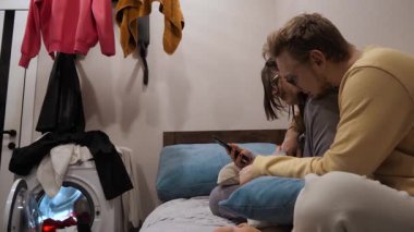 Çift, rahat bir yatakta akıllı bir telefonla oturmaktan zevk alıyor. Dijital dünyada rahat bir ev ortamında, rahat yaşam tarzlarını yansıtan bir çamaşır makinesiyle.