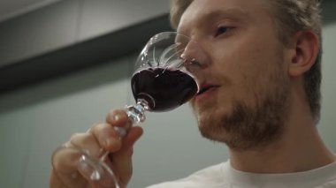 Düşünceli genç adam modern mutfakta bir bardak kırmızı şarabın tadını çıkarıyor. Rahatlama ve zevk anını yansıtıyor. Kapatın.