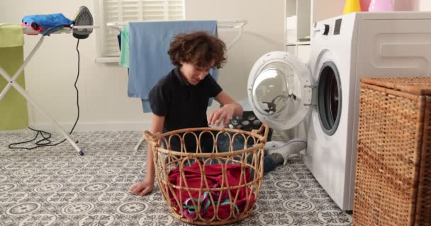 一个穿着牛仔裤和衬衫 面带微笑的少年坐在一台家用洗衣机前 他把脏衣服装入洗衣机 一间明亮 通风的洗衣房 — 图库视频影像