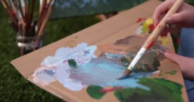 Bir kadın sanatçının elinin yakın plan resmi, ilkbahar zamanı yeşil bir alanda bir tuval manzarası çiziyor. Bahçede resim atölyesi. Sanat eğitimi kavramı. Açık hava eğlence aktiviteleri