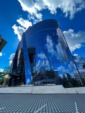 Mavi gökyüzünü ve bulutları yansıtan modern cam gökdelen.