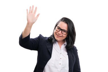 Adult entrepreneur woman with friendly expression smiling wearing spectacles and formal suit waving hand as hello gesture isolated on white studio background