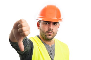 Ciddi inşaatçı, sinirli görünüşlü, turuncu kasklı ve yelekli bir erkek model. Beyaz stüdyo arka planında izole edilmiş, beğenilmeyen baş parmak hareketi gösteriyor.