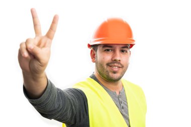 Dost canlısı yetişkin inşaatçı gülümseyen ve beyaz stüdyo arka planında izole edilmiş floresan yelek ve turuncu şapka giyen parmaklarıyla barış ya da zafer işareti gösteren.