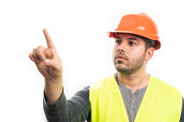 Güvenlik kaskı ve ciddi ifade içeren floresan yelek giyen erkek müteahhit, beyaz ekranda izole edilmiş şeffaf ekran görüntüleme boşluğuna dokunmak için işaret parmağı kullanıyor.
