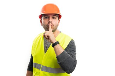 İş yerindeki yetişkin müteahhit, güvenlik kaskı floresan yelek giyiyor. Ağız üzerinde işaret parmağı olan sessiz bir jest yapıyor.