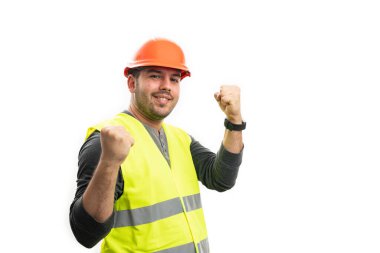 Turuncu güvenlik kasketi ve sarı floresan yelek giyen erkek müteahhit, kutlama yaparken gülümsüyor. Yumruk hareketi yapıyor ve boş telif alanı beyaz üzerinde izole ediliyor.