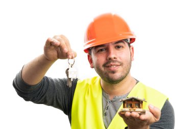 Turuncu şapkalı, sarı yelekli, gülümseyen yetişkin müteahhit beyaz arka planda izole edilmiş ev anahtarlarını elinde tutan minyatür ahşap evi sunuyor.