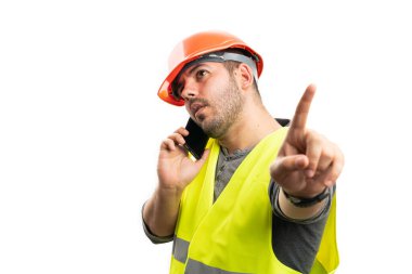 Erkek inşaat işçisi telefonda ciddi bir ifadeyle konuşuyor. Beyaz arka planda izole edilmiş işaret parmağı kullanarak bekleme hareketi yapıyor.