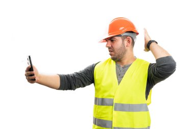 İş yerindeki müteahhit, beyaz stüdyo arka planında izole edilmiş akıllı telefon kullanarak video görüşmesinde turuncu güvenlik kaskı ve floresan sarı yelek giyiyor.