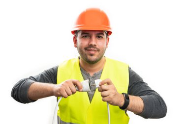 Gülümseyen elektrikçi turuncu kasketli güvenlik ekipmanı takıyor uzatma kablosuna elektrik prizi takıyor beyaz stüdyo arka planında izole edilmiş.