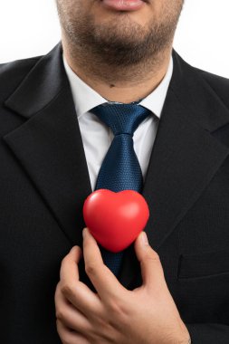 Yetişkin iş adamı, beyaz stüdyo arka planında izole edilmiş sembol gibi kırmızı kalbi göğsüne yakın tutan şık ofis kıyafeti giyiyor.