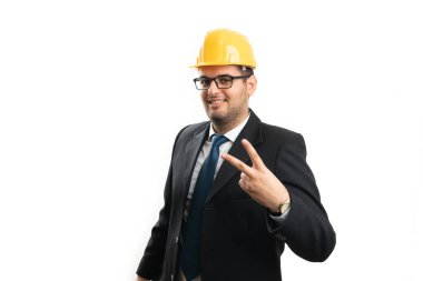 Yetişkin bir mimar ofis kıyafeti ve güvenlik kaskı giyiyor. Stüdyo arka planında parmakları izole bir şekilde barış ya da zafer işareti gösteriyor.