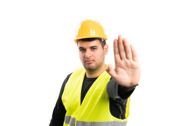 Sarı güvenlik kaskı ve yelek giyen ciddi bir inşaat işçisi, beyaz stüdyo arka planında avuç içi izole edilmiş, bekleme hareketi yapıyor.