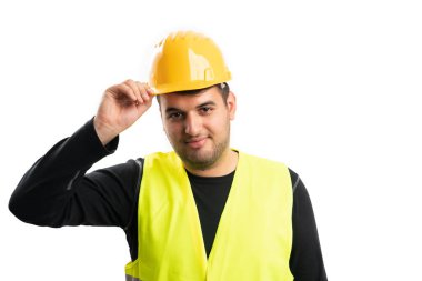 Dost canlısı inşaatçı, floresan iş yeleği giyiyor. Beyaz arka planda izole edilmiş sarı güvenlik şapkasına dokunarak selam veriyor.