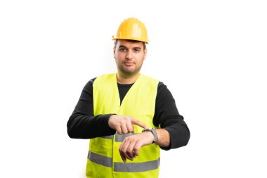 Ciddi bir ifadeyle yetişkin bir erkek müteahhit, beyaz stüdyo arka planında izole edilmiş zaman tutma kavramı olarak inşaat kıyafeti giymiş işaret parmağıyla kol saatine işaret ediyor.