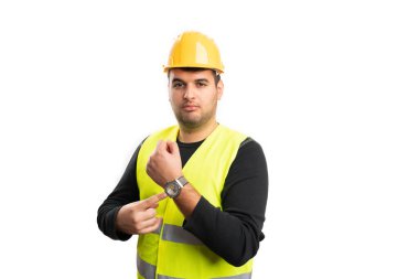 Güvenlik ekipmanı yeleği giyen ciddi bir erkek inşaat işçisi ve beyaz arka planda izole edilmiş zaman tutma kavramı olarak işaret parmağıyla kol saatine işaret eden bir şapka takıyor.