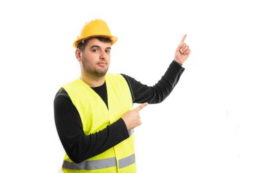 Sarı kasketli ve floresan yelek giyen erkek inşaat işçisi beyaz stüdyo arka planında izole edilmiş reklamlar için boş telif alanını işaret ediyor.