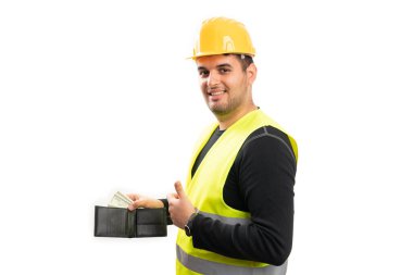 Erkek inşaat işçisi floresan yelek ve sarı güvenlik kaskı takıyor. Deri cüzdanı ve nakit para gösteriyor. Başparmağını kaldırıyor.