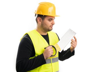 Sarı koruyucu kask ve floresan yelek giyen erkek inşaat işçisi, beyaz stüdyo arka planında izole edilmiş parmak kullanarak el testeresine dokunuyor.