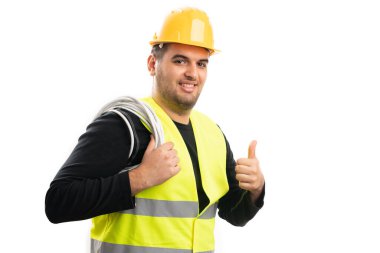 İş kıyafetleri içinde gülümseyen erkek müteahhit omzunda uzatma kablosu tutuyor. Beyaz stüdyo arka planında izole edilmiş bir jest gibi baş parmağını gösteriyor.