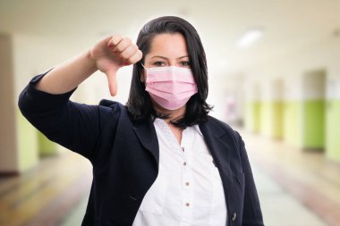 Corporate businesswoman making thumb-down disagree dislike gesture wearing pink disposable mask for covid19 prevention as pandemic concept with building background clipart