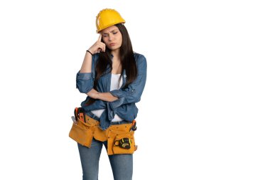 Sarı kasketli ve kot gömlekli genç bir kadın, alet kemeri takıyor ve düşünceli bir ifadeyle ayakta duruyor. Bir inşaat ya da DIY setinde konsantrasyon ve karar verme gibi bir sorunu çözmeye çalışıyormuş gibi alnını ovalıyor.
