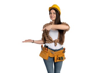Sarı bir şapka ve alet kemeri takan gülümseyen kadın inşaat işçisi, sanki bir ürün ya da fikir sunuyormuş gibi avucunun içini işaret ediyor. Beyaz bir arkaplanda duruyor.