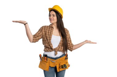 Kendine güvenen bir kadın inşaat işçisi sarı bir şapka takıyor ve iki eliyle de kemer takıyor, bir şey gösteriyor. Gülümser ve beyaz bir arkaplanda durur..