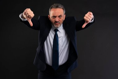 Takım elbiseli olgun bir adam, iki baş parmağı aşağıda ve kızgın bir ifadeyle güçlü bir hoşnutsuzluk gösteriyor. İş ortamında reddedilmeyi, hayal kırıklığını ya da anlaşmazlığı iletiyor..