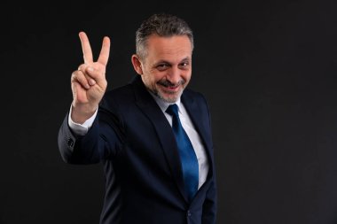 Resmi takım elbiseli kendine güvenen, gülümseyen ve barış ya da zafer jesti olarak iki parmağını kaldırıp karanlık bir arka plana karşı duran olgun bir adam..